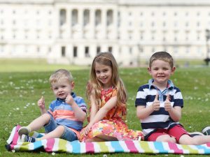 Two small boys and a girl sitting on striped map on grass in front of Parliament Buildings Stormont